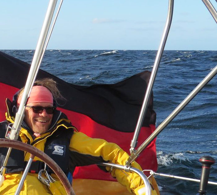 Skipper mit Ölzeug und Weste vor wehender Bundesflagge am Heck. Segeln raumschots im Herbst bei Sonne auf der Ostsee.